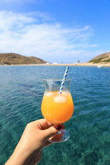 Summer drink cocktail of fresh orange juice with ice in the woman hand on the seascape background of Kolona beach Kythnos island Cyclades Greece.
