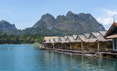 Fototapeta na wymiar Wooden Thai traditional floating houses on a lake with mountains and rain forest in the background during a sunny day at Ratchaprapha Dam at Khao Sok National Park, Thailand
