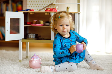 Adorable cute little toddler girl playing with toy kitchen Happy healthy baby child having fun with role game, playing with crockery teapot at home or nursery. Active daughter with toy.