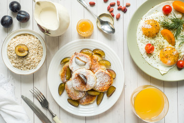 Concept of Breakfast. Fried eggs, cottage cheese pancakes, plums and oatmeal with milk, orange juice on the table