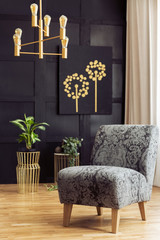 Patterned armchair in dark living room interior with chandelier above plants and poster. Real photo
