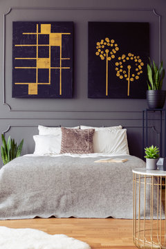 Black and gold posters on grey wall above bed with pillows in bedroom interior with plants. Real photo