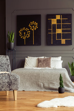 Patterned armchair next to bed in grey bedroom interior with black posters and plants. Real photo