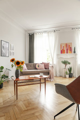 Wooden coffee table with fresh sunflowers, open book and tea cup in real photo of bright living room interior with window with drapes