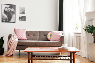 Real photo of couch with pastel pink cushions and blanket standing in white sitting room interior with wooden table with book and coffee cup, window with curtains and posters on wall