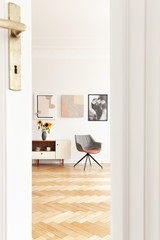 Real photo through the door of bright living room interior with grey chair, cupboard with sunflowers, three posters and herringbone parquet
