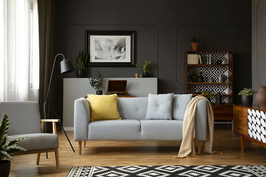 Real photo of grey sofa with pillows standing in dark living room interior with molding on wall, metal lamp, vintage cupboards and window with drapes