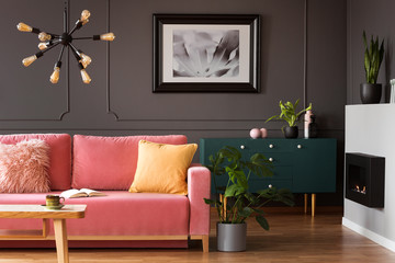 Real photo of powder pink sofa with open book standing in dark living room interior with wainscoting wall, eco fireplace and fresh plants