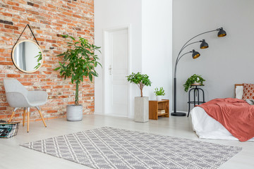 Mirror on red brick wall above grey armchair in apartment interior with plants and bed. Real photo