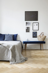 Grey blanket on bed with blue pillows in white bedroom interior with posters above bench. Real photo