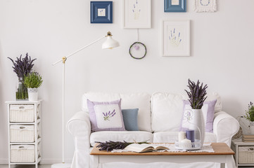 Lavender flowers on cabinet next to lamp and white sofa in living room interior with table and posters. Real photo