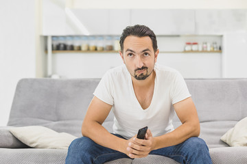 Nothing interesting to watch. Handsome young man holding remote control and looking bored while watching TV on the couch at home