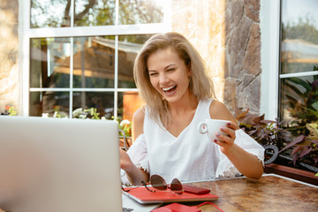 Attractive blonde businesswoman laughing while looking at laptop screen, woman drinking a cup of tea or coffee, during spending time in cafe. Smartphone, sunglasses and notebook are on the table.