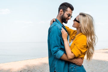 side view of couple in sunglasses hugging and looking at each other near ocean