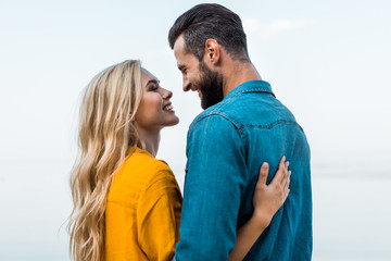 Fototapeta premium side view of smiling couple hugging and looking at each other against blue sky
