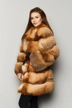 Fashionable and stylish brunette model girl with perfect makeup and blue-grey eyes, in luxury fur coat ginger colour, smiling and posing at studio, isolated