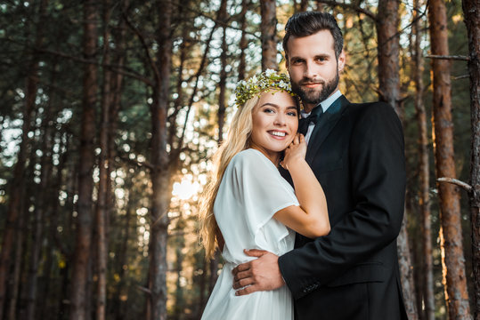low angle view of smiling wedding couple hugging and looking at camera during sunset in forest