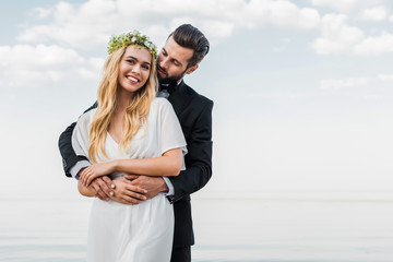 handsome groom in suit hugging attractive bride on beach
