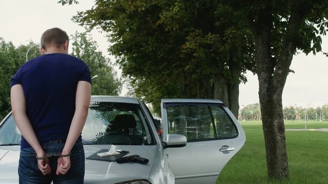 detained burglar in handcuffs stands near a stolen car