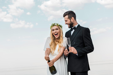 happy bride in white dress opening champagne bottle on beach
