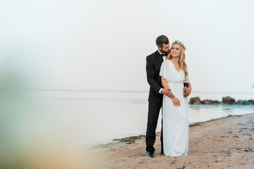 groom in suit hugging attractive bride in white dress on beach