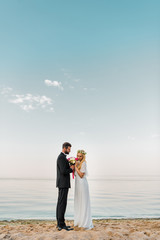 side view of wedding couple standing with bouquet on sandy beach, bride sniffing roses