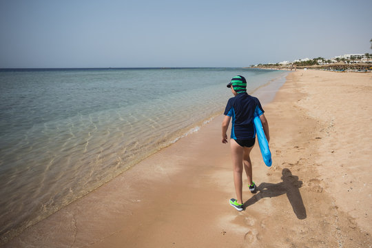 Cute white tanned kid dressed in swimsuit, cap and aqua shoes having fun at summer sandy beach using blue inflatable surf board. Boy walking alone with his back to camera. Horizontal color photography