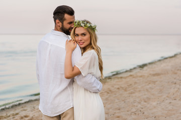groom hugging attractive bride in wreath and she looking at camera on beach