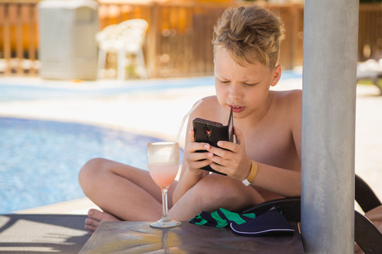 Funny child with serious expression on cute face drinking cocktail and usimg modern mobile cellphone for entertainment. Kid sits on beach chair at poolside area of tropical hotel resort. 