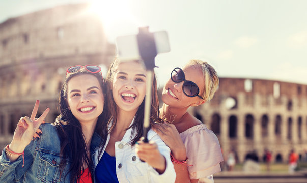summer vacation, holidays, travel, technology and people concept- group of smiling young women taking picture with smartphone on selfie stick over coliseum in rome background
