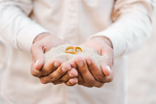 cropped image of groom holding wedding golden rings with sand in hands on beach