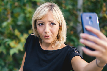 Blonde young adult woman taking selfies with the smartphone in a public park