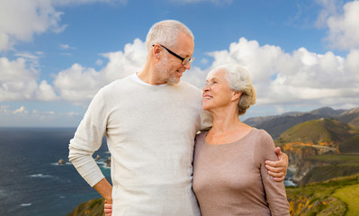 old age, tourism, travel and people concept - happy senior couple hugging over bixby creek bridge on big sur coast of california background