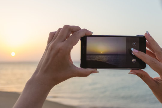 Happy european woman taking photo of sunrise or sunset sea using digital camera of smartphone during summer vacation. Closeup color photography of female hands making pictures of landscape.