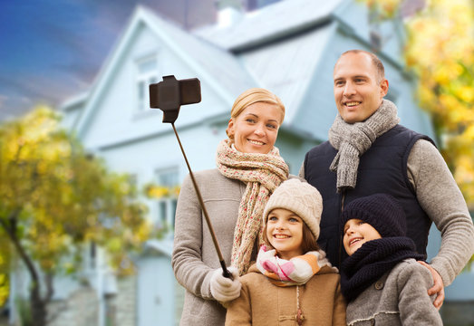 family, technology and real estate concept - happy mother, father, daughter and son taking picture by smartphone on selfie stick over living house background outdoors in autumn