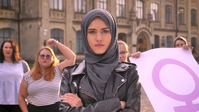 Group of various caucasian feminists with cute girl in hijab in center attracting attention with pink flag and voting outdoor