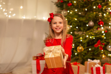 Fototapeta na wymiar christmas, holidays and childhood concept - smiling girl in red dress with gift box at home
