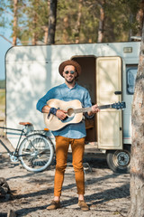 male guitarist playing on acoustic guitar near campervan with bike