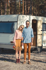 hippie couple in wreaths and sunglasses holding hands and posing near camper van