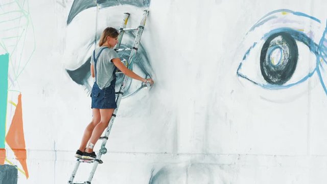 Beautiful girl making graffiti of big female face with aerosol spray on urban street wall. She standing on ladder. Creative art. Talented student in denim overalls drawing picture