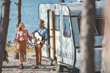 hippie girlfriend dancing while boyfriend playing guitar near trailer in nature