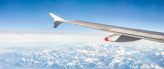 Panoramic image of a plane wing over snowy alpine mountains on a sunny day