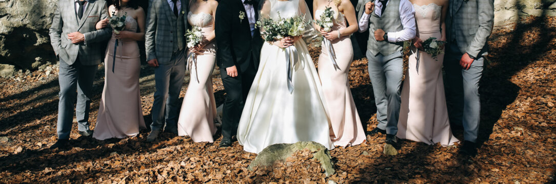 Wedding couple is with their friends in the autumn wood. Best man in stylish elegant checkered suits and bridesmaids in powder pink pastel dresses are standing near the bride and groom outdoors.