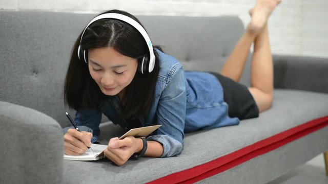 Happy young woman lying on the sofa drawing something in note book and listening music