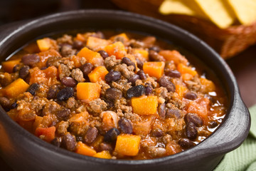 Homemade chili con carne with mincemeat, red and black beans, tomato sauce and pumpkin in rustic bowl with tortilla chips in the back (Selective Focus, Focus in the middle of the dish)