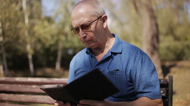 An elderly man with glasses reading an e-book on a Park bench