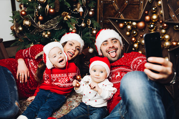Portrait of happy funny parents and their two children in red sweaters and hats making selfie via...
