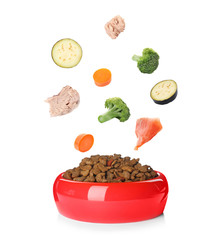 Fresh ingredients falling into bowl with dry pet food for cats and dogs on white background