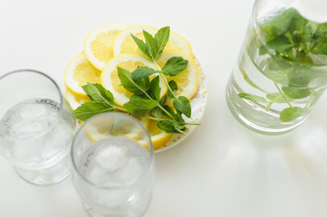 Obraz na płótnie Canvas Top view of two glasses with ice cubes, plate with lemon slices and mint leaves and also bottle of water, all on white table.