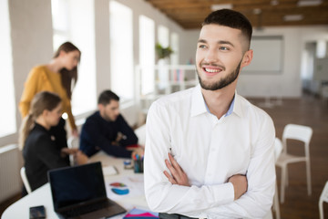 Young smiling man with beard in white shirt happily looking aside with folded hands while spending time in office with colleagues on background